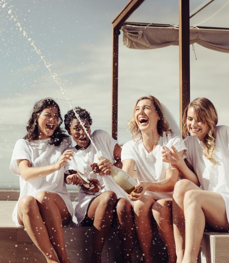Cheerful bride and bridesmaids celebrating hen party with champagne while sitting on rooftop. Girls having a great time at the hen party.